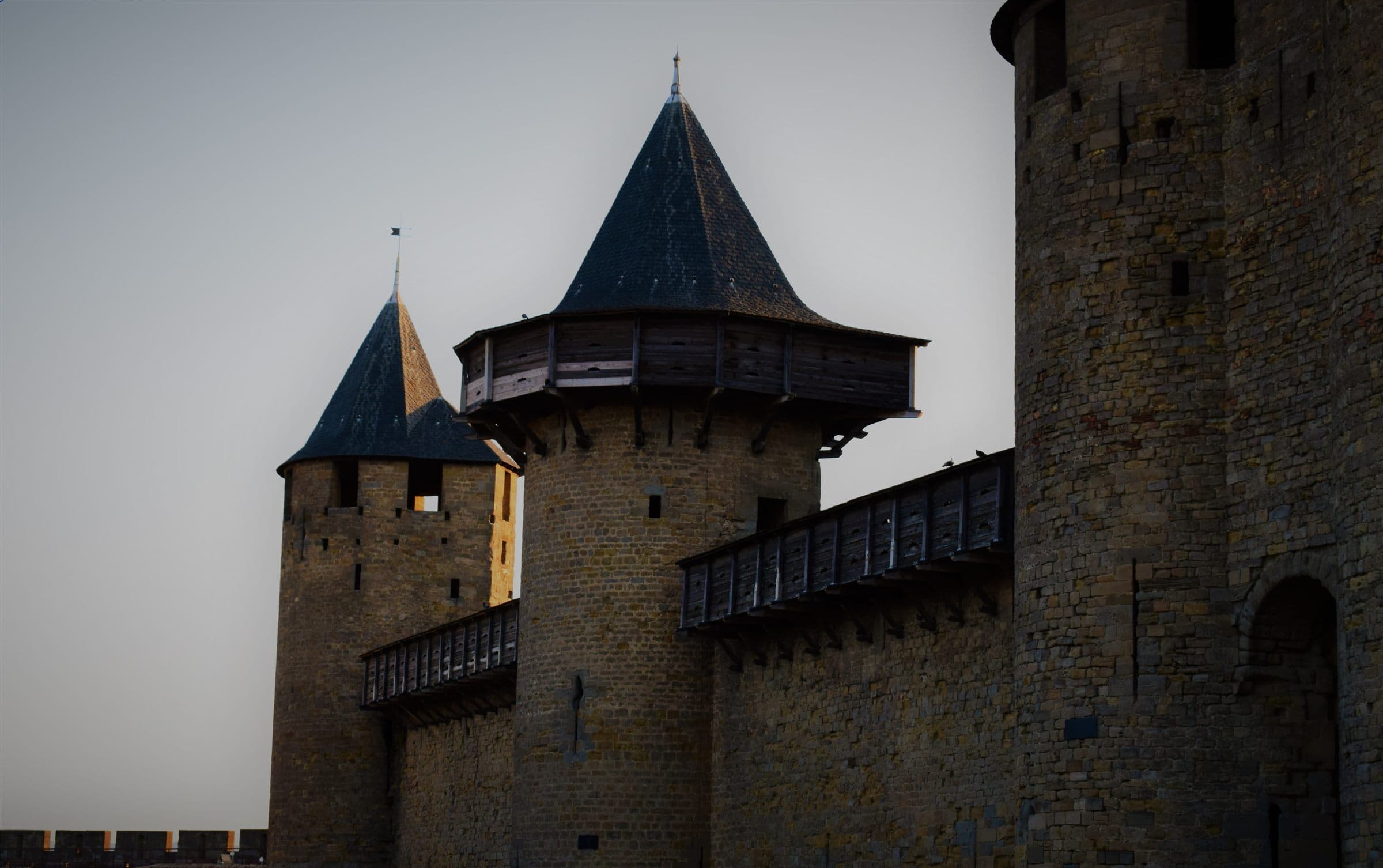 Stone towers and timber hoardings of the Château Comtal at dusk, the last sun catching one turret